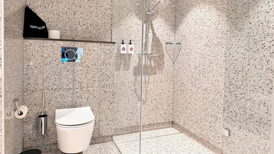 A modern bathroom features a wall-mounted toilet and an adjacent shower area with a rainfall showerhead. Grey patterned tiles cover the walls. A shelf holds a black bag labeled "Hairdryer."
