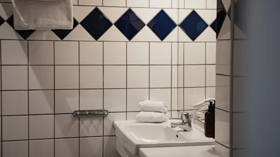 Et rent badeværelse med en hvid vask, foldede håndklæder og en sæbedispenser. Væggene er flisebelagt med hvide firkanter og et blåt diamantmønster.