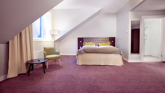 Et pent ordnet hotellrom med en seng, stoler, og et dusjrom, preget av skrå vegger og dempet belysning.