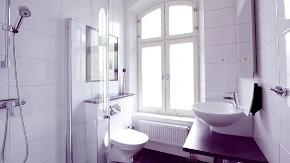 A bright, tiled bathroom with a glass-enclosed shower, toilet, and modern basin on a dark countertop, lit by natural light from arched windows.
