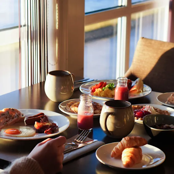 Frokosttallerkener arrangert på et solfylt spisebord inkluderer egg, pølser, croissanter, frukt og juice. En hånd holder et redskap ved siden av krus og glassflasker i et rom med store vinduer.