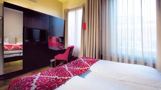 Hotellrom med oppredd seng med røde blomstermønstre, en veggmontert TV og lyse gardiner; moderne innredning.