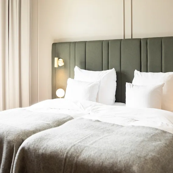A bright hotel room with two comfortable beds, a dark green headboard, and a large window with light curtains.