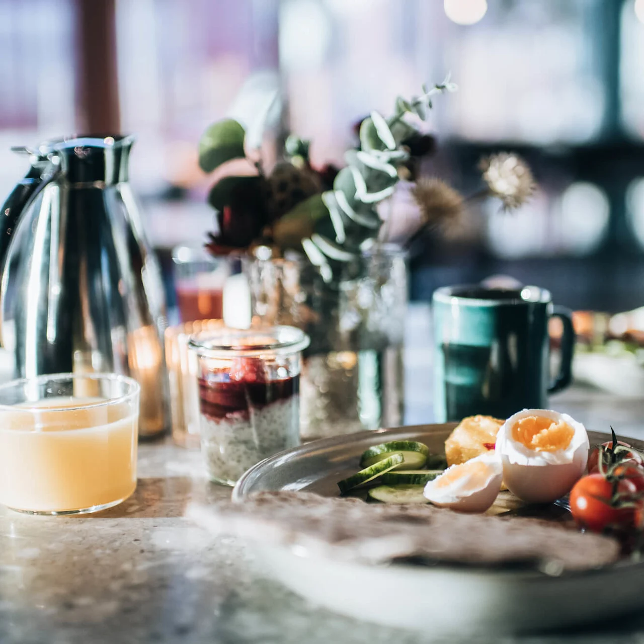 A breakfast plate with sliced cucumber, halved boiled eggs, and cherry tomatoes sits on a table, accompanied by a glass of juice and a carafe in a softly lit kitchen.