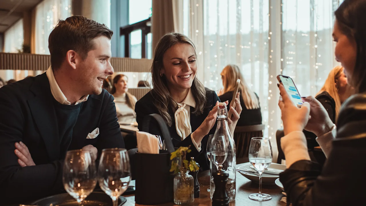 Three people enjoying a meal at a restaurant, with one person taking a photo or showing something on their phone.