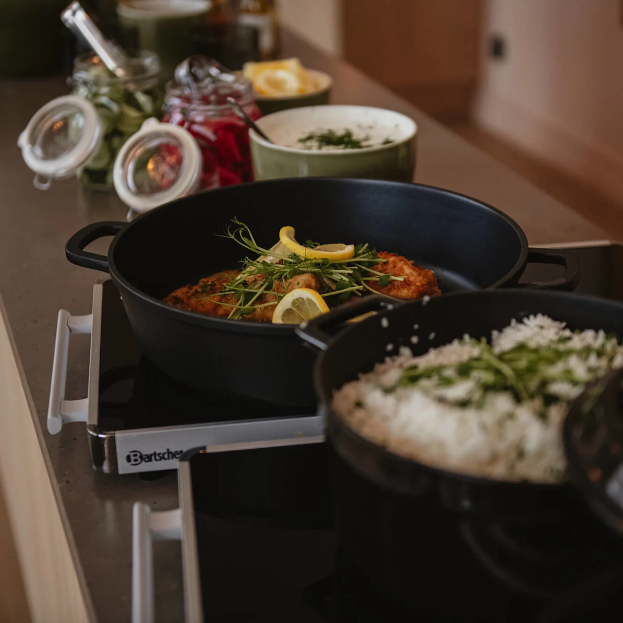 English: Black pots on a kitchen counter, one containing a breaded dish garnished with lemon and herbs, another with rice topped with green sprigs, surrounded by jars and bowls. "Bartscher" text visible.