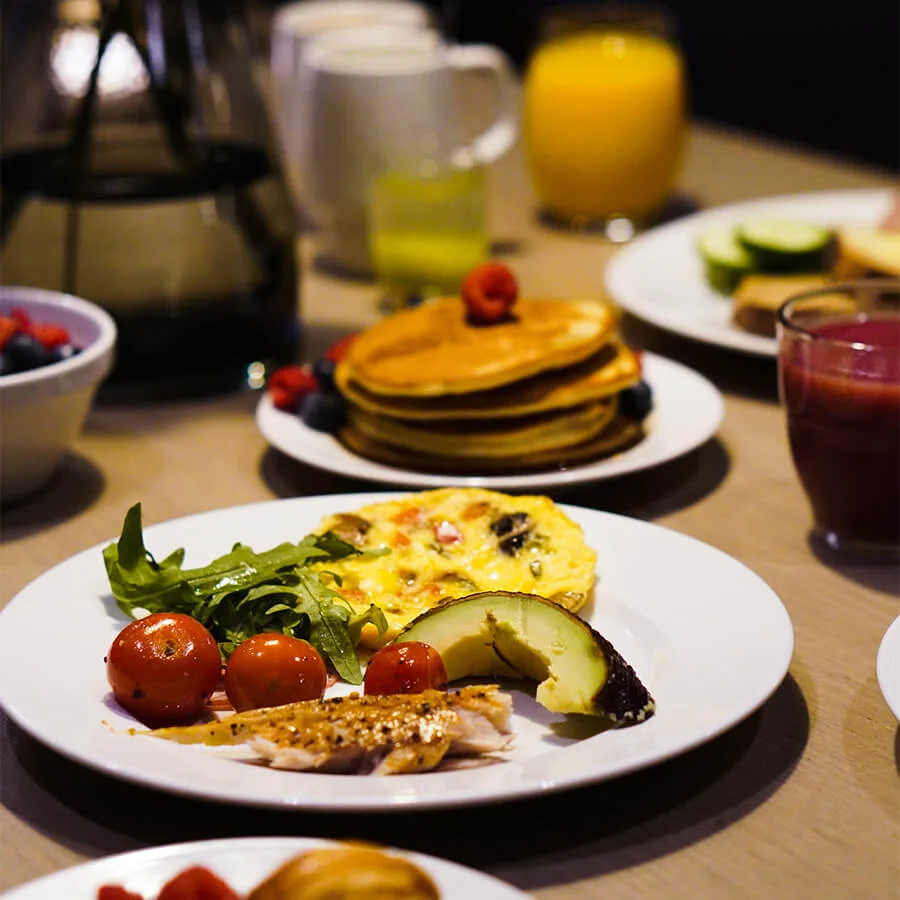 En rekke frokostretter, inkludert pannekaker, omelett, ruccola, tomater og avocado på tallerkener, med drikker som appelsinjuice og kaffe på et trebord.