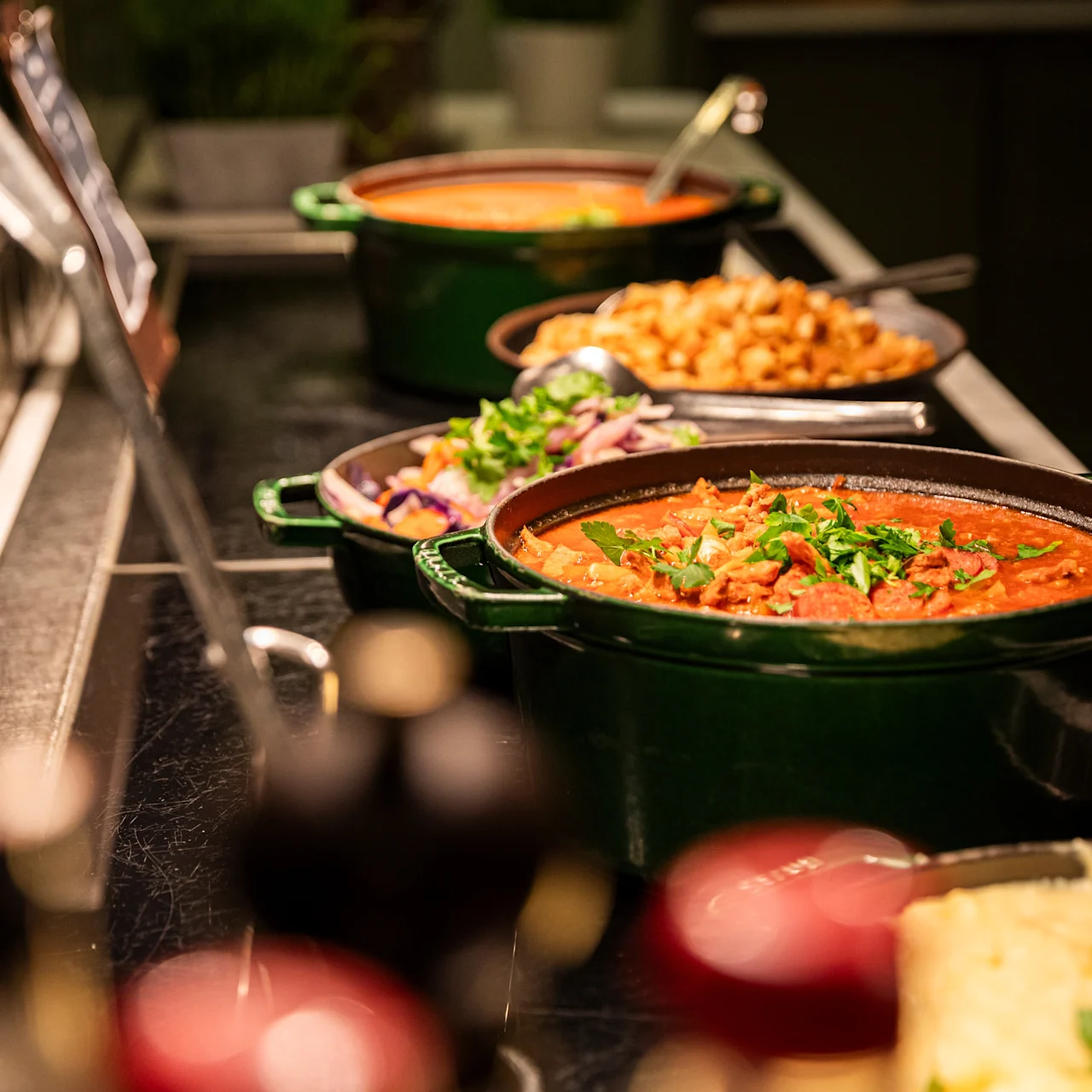 Green pot holds stew topped with herbs, sitting on a buffet table alongside other dishes and utensils, under warm lighting.