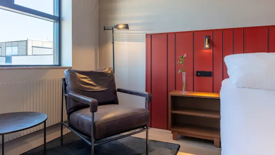 A leather armchair beside a window in a modern room with a red accent wall, bed, and wooden furniture.