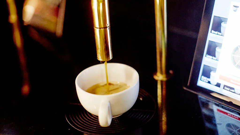 Kaffekop fyldes med espresso fra en gylden dispenser, omgivet af en dæmpet belyst miljø. En digital skærm viser drikkevaremuligheder. Tekst: "Cold milk".