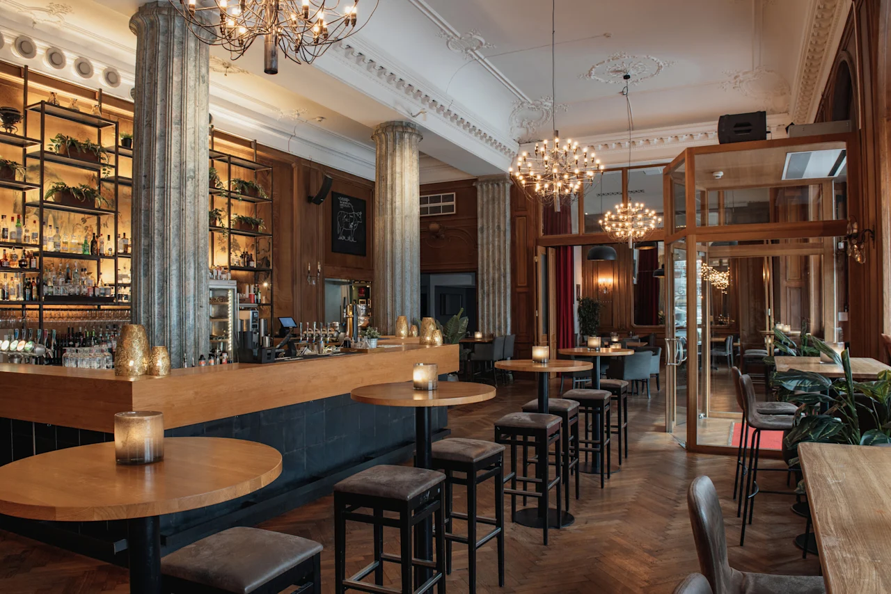 Elegant hotellbar på Home Hotel Grand, Helsingborg, med en lång trädisk, höga bord och stiliga ljuskronor.