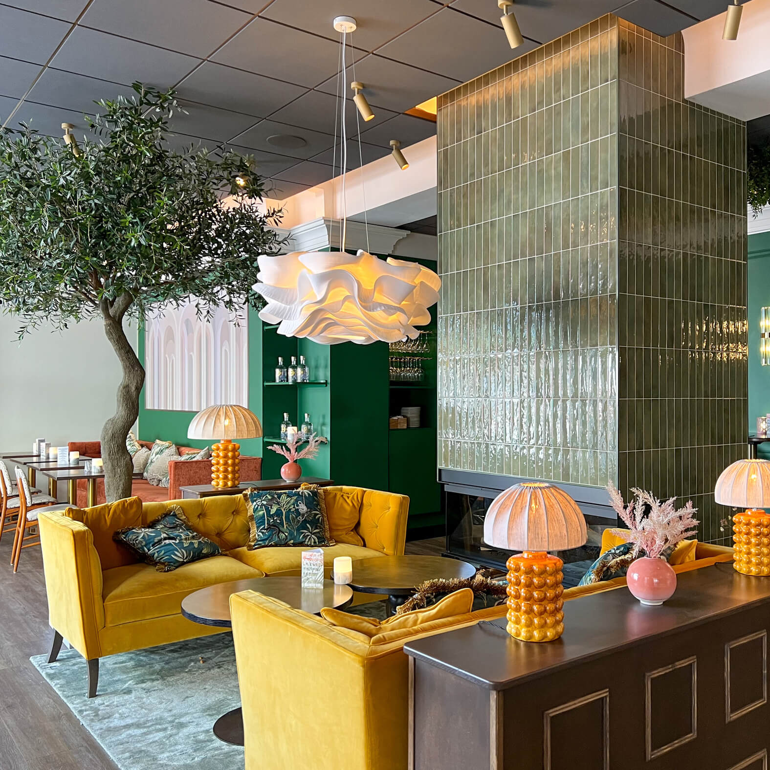 A vibrant, eclectic dining area featuring mustard yellow sofas, textured glass partitions, and decorative lighting, surrounded by greenery accents and chic decor.