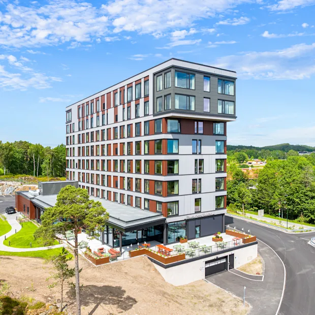 A modern hotel building with a unique design, surrounded by lush green trees and a winding road under a blue sky.