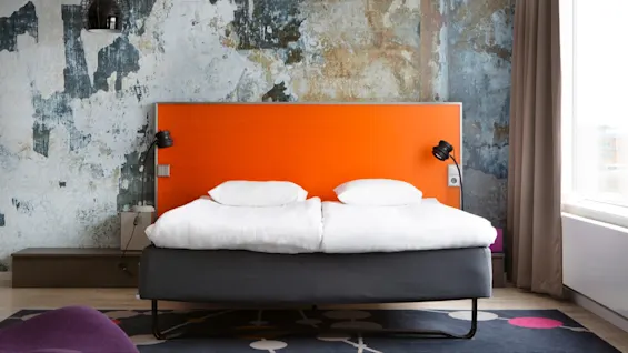En seng med orange sengegavl står mod en struktureret, slidt væg. To hvide puder ligger på den, omgivet af moderne lamper og et stort vindue med gardin.