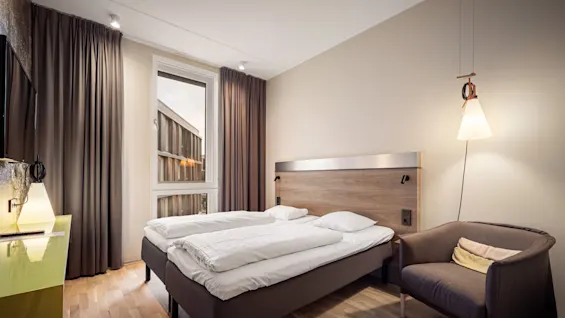 A neatly made bed with white linens is placed in a modern hotel room, featuring a large window, curtains, a lamp, and an armchair on a wooden floor.