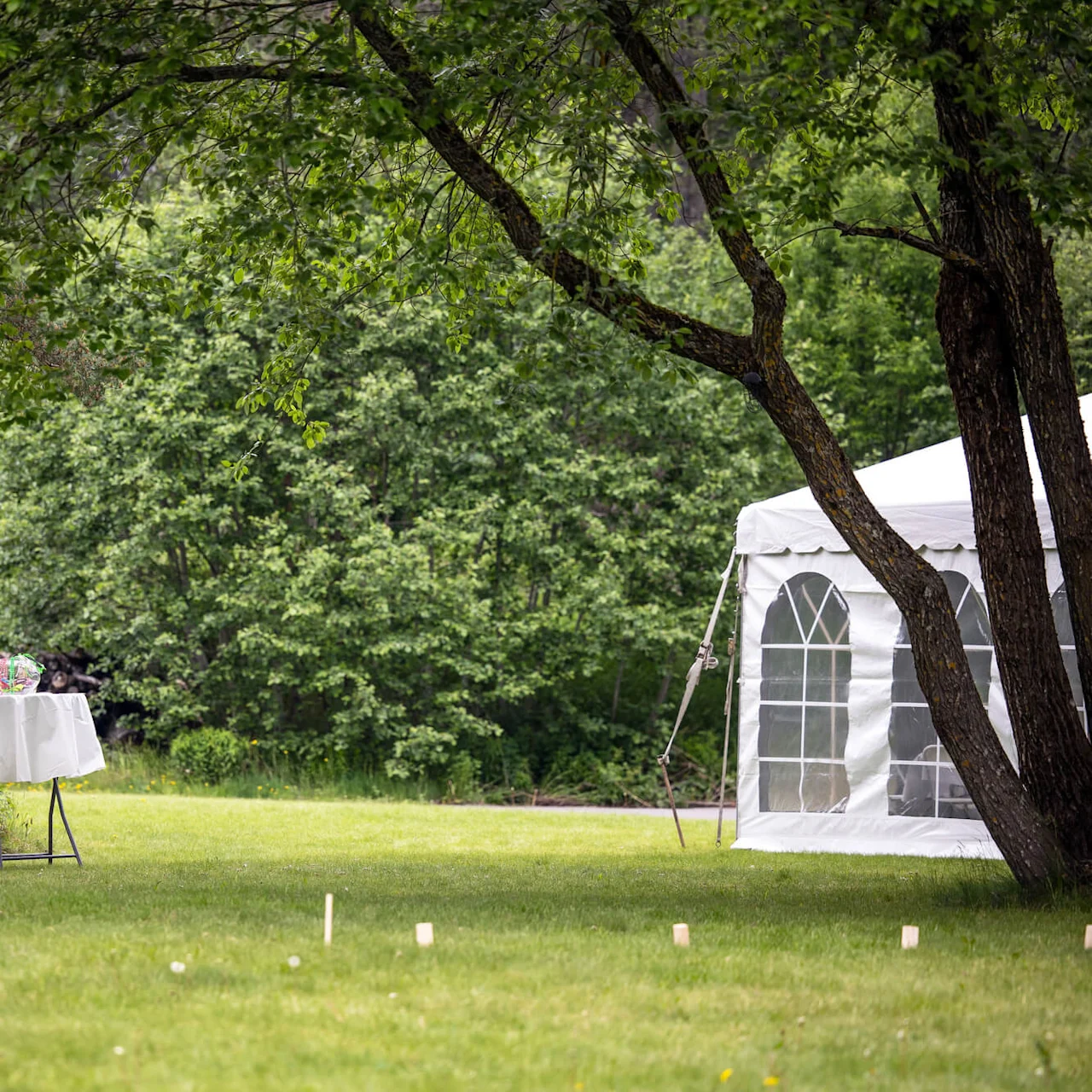 A table with a white cloth holds a glass jar in a lush garden beside a floral wall and a white tent. Tall trees and green bushes surround the area.