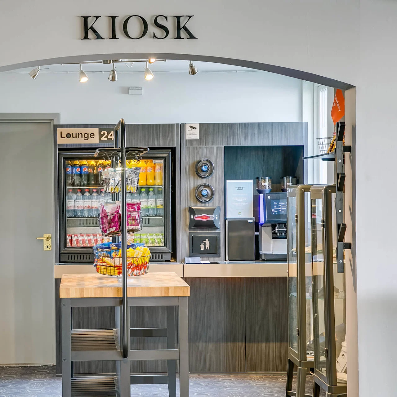 Kiosk med drikkevarer og snacks fremvist i moderne hotelomgivelser. Skilte lyder "QUALITY HOTEL," "KIOSK," og "Lounge 24." En automat og kaffestation er til stede.