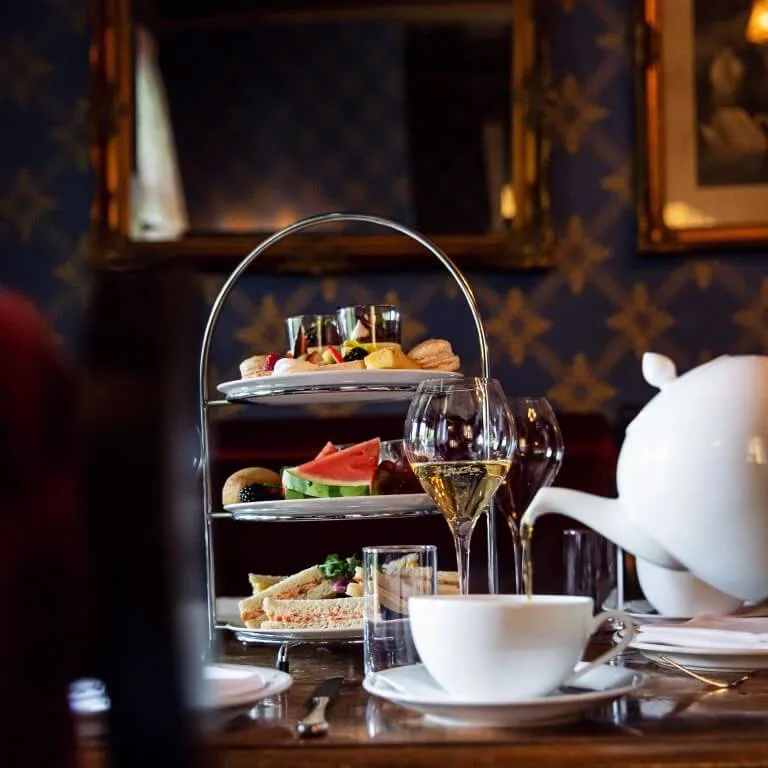 Afternoon tea set on a wooden table with a two-tier tray of sandwiches and pastries. A teapot, teacup, and wine glass are arranged elegantly in a cozy, decorated room.