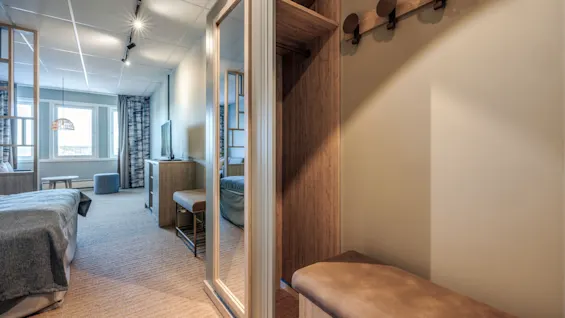 A cozy hotel room features a bed, a TV on a stand, and a hallway with a bench and coat hooks. Soft lighting and large window complete the relaxing ambiance.