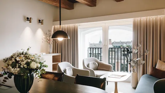 Elegant hotellsvit med modern sittgrupp, takbjälkar i trä och stora fönster med utsikt över staden.