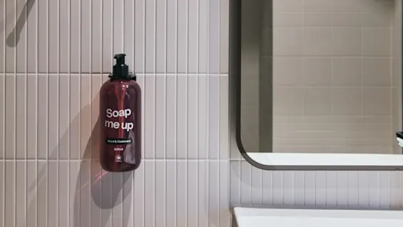 En badrumsscen med en tvåldispenser märkt 'Soap me up Wood & Cashmere 500ml' på en kaklad vägg bredvid en spegel och ett handfat.