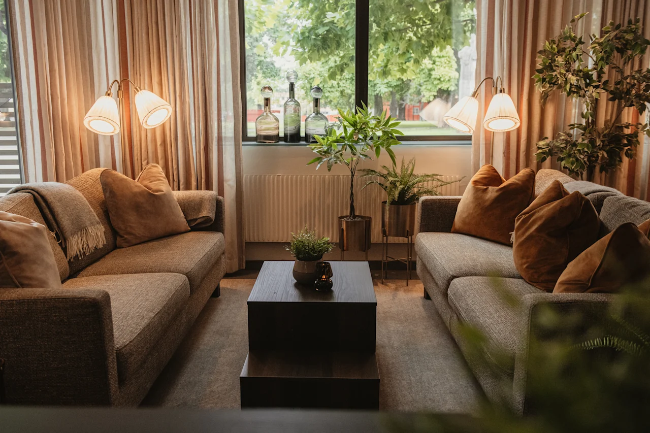 A cozy living room with two sofas, a coffee table, and plants, bathed in natural light from a large window overlooking greenery.