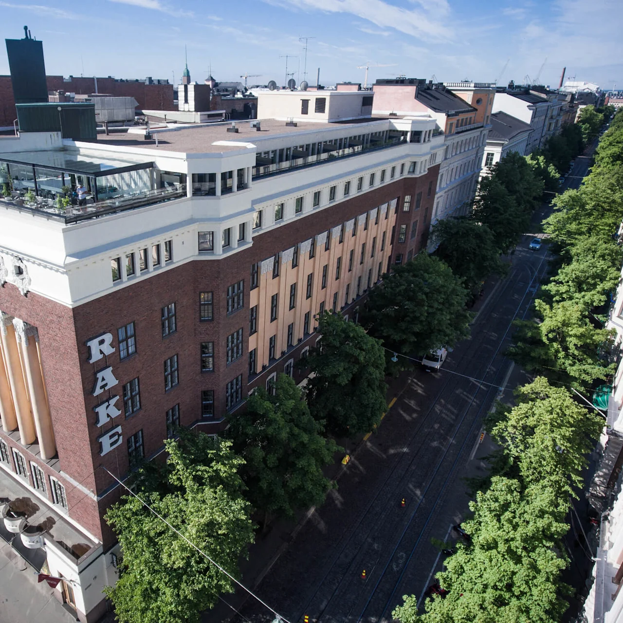 Suuri tiilirakennus, jossa lukee "RAKE", seisoo kulmassa. Siinä on kattoterassi, jossa on pöytiä ja tuoleja, ympäröitynä puiden reunustamalla tiellä kaupunkiympäristössä.