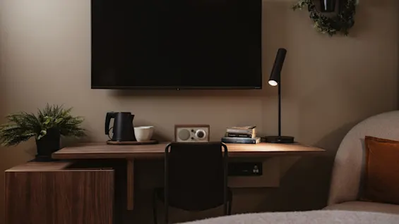 A wall-mounted TV overlooks a wooden desk with a lamp, kettle, books, and plant; a cozy room setting.