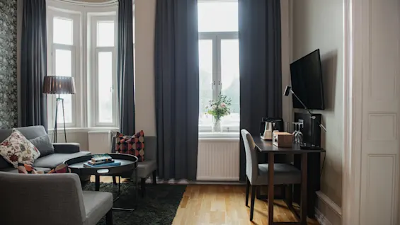 En hyggelig stue med en grå sofa og lænestol med farverige puder, placeret rundt om et sofabord. I nærheden står et skrivebord med et tv vendt mod et vindue med mørke gardiner.