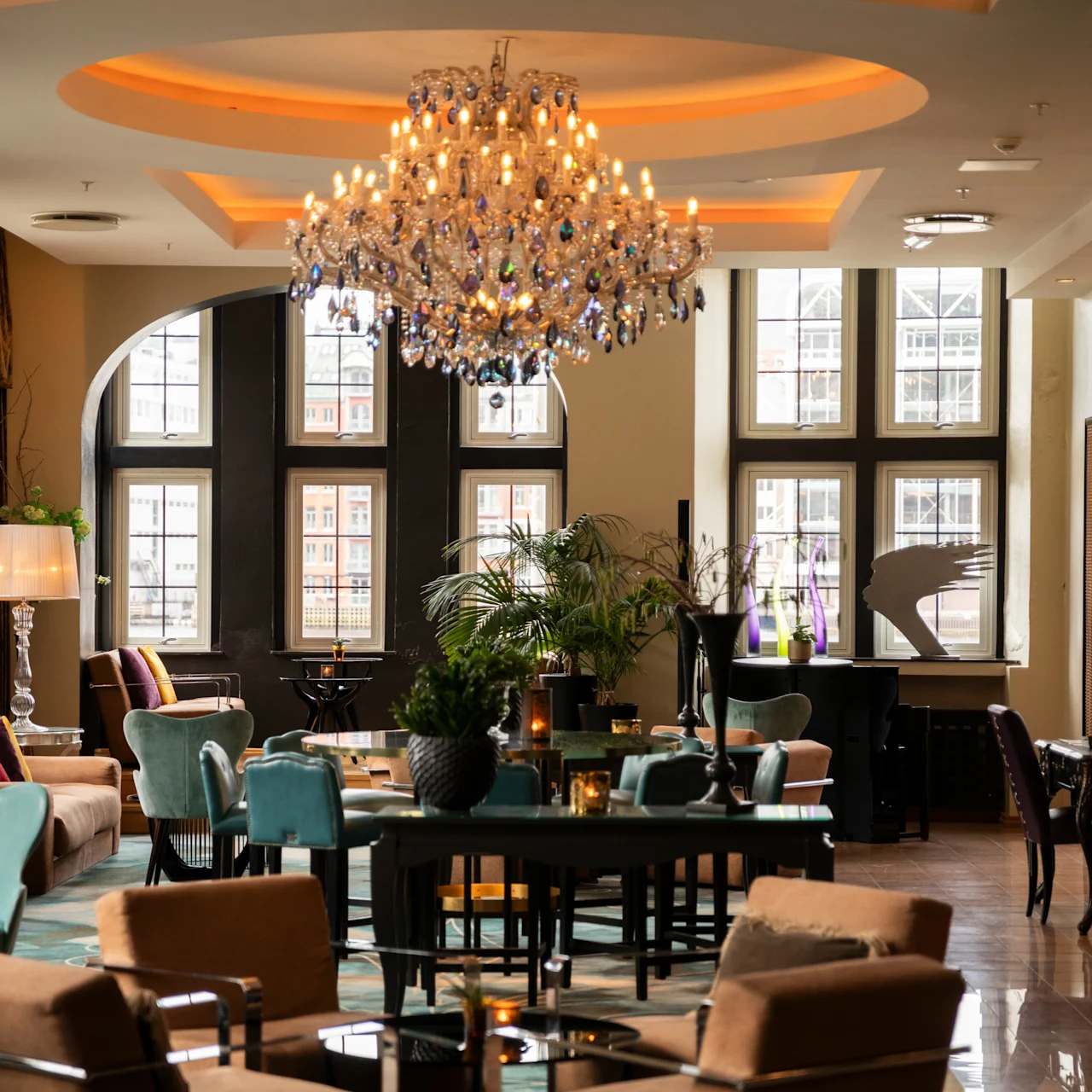 Luxurious chandelier illuminating a cozy lounge adorned with sofas, chairs, and tables; surrounded by large windows and elegant decor, creating an upscale, inviting atmosphere.