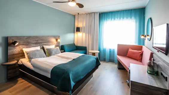A modern hotel room features a neatly made bed with teal bedding, side tables, and a ceiling fan, surrounded by a sofa and bright window with curtains.