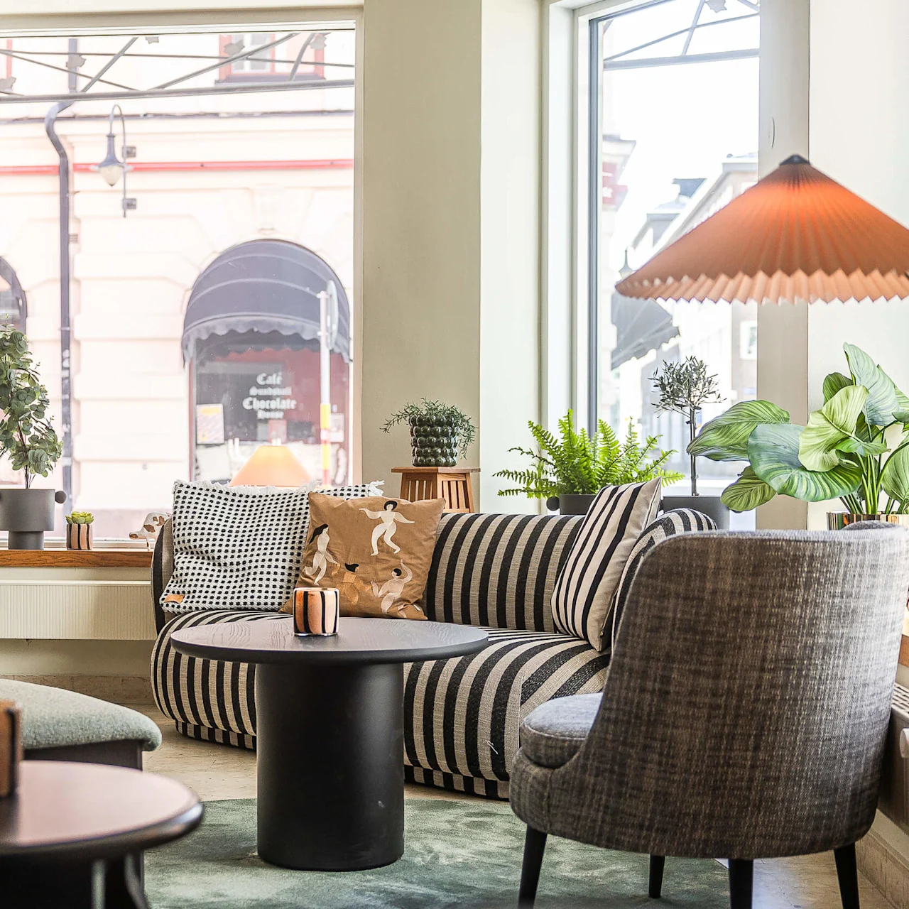 En mysig interiör med en randig soffa, växter, ett svart bord och en lampa, stort fönster med utsikt över gatan och ett café mittemot.
