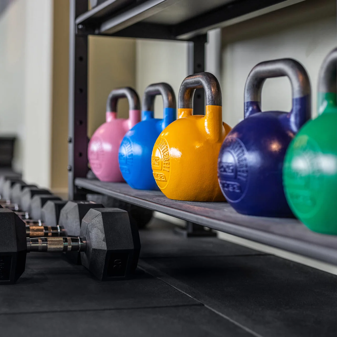 Fargerike kettlebells på en hylle, sammen med rader av svarte manualer på gulvet, i et treningsstudio. Matt gulv og dempede vegger gir en moderne, funksjonell bakgrunn.