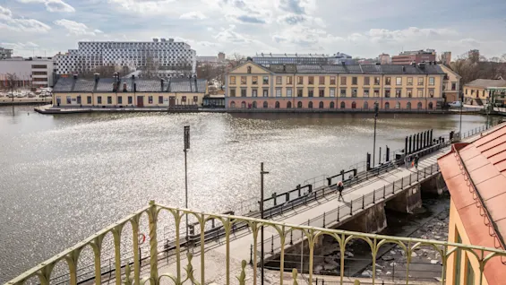 En stor gul byggnad med bågformade fönster står vid en skimrande flod under en delvis molnig himmel. Människor promenerar längs en närbelägen bro, omgivna av en stadsmiljö.