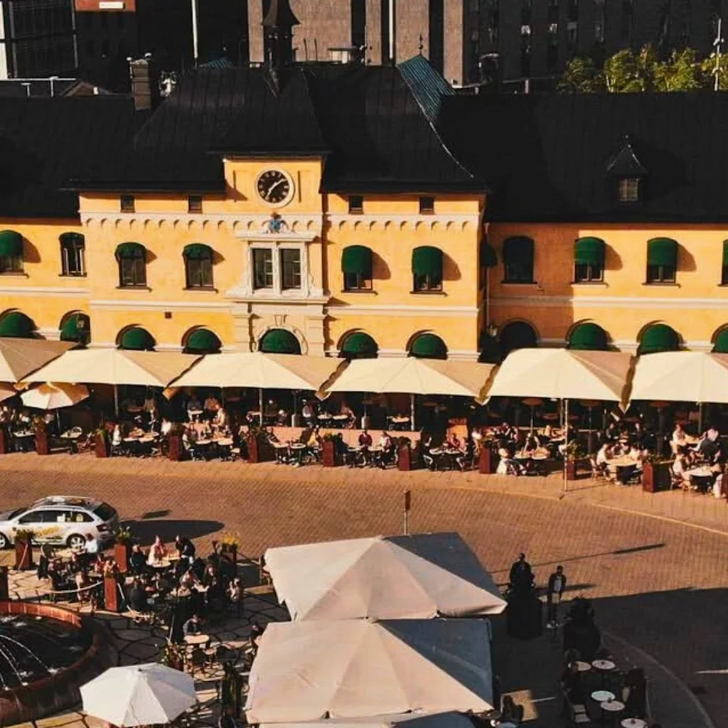 A vibrant outdoor dining area in front of a yellow building, with many people enjoying meals under umbrellas.