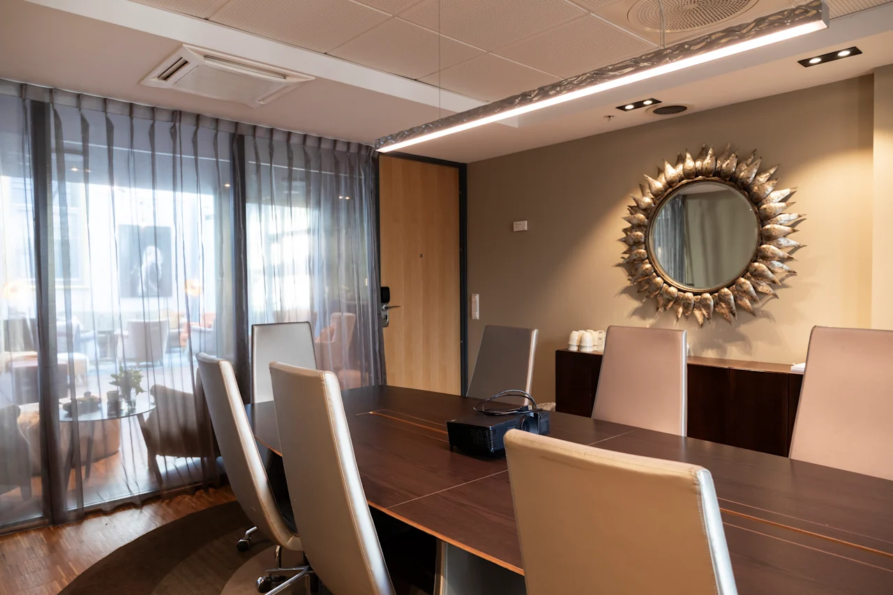 Ett konferensbord omgivet av stolar står i ett mötesrum med en dekorativ solspegel på väggen, skira gardiner och mjuk belysning som skapar en professionell atmosfär.