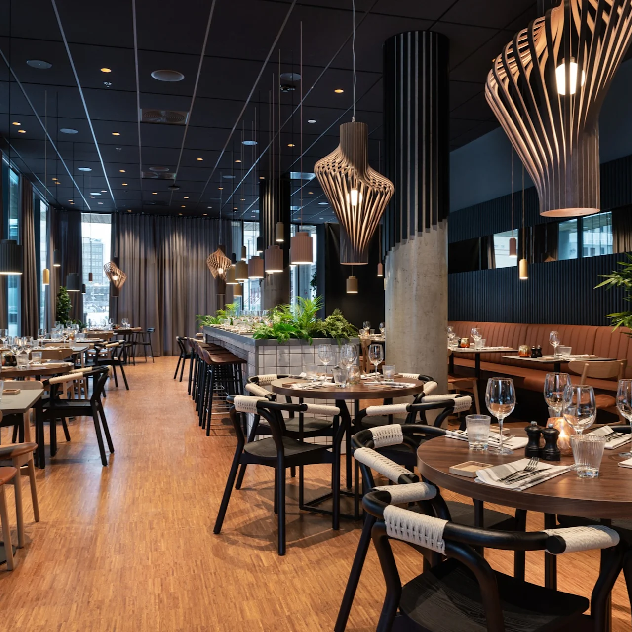 Bordene er elegant dekket med glass og bestikk i en moderne restaurant, med tremøbler, stilige pendellamper og grønne planter som skaper en varm og innbydende atmosfære.
