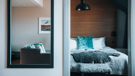 Et hyggeligt soveværelse med turkise accenter, som har en seng med puder og et tæppe, ved siden af et spejl, der reflekterer en sofa og indrammet flyfoto på en beige væg.