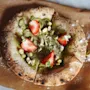 A round pastry topped with green spread, sliced strawberries, and chopped pistachios rests on brown parchment paper, placed on a marble surface.