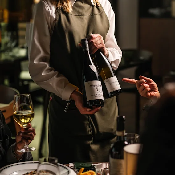 En servitör i förkläde presenterar två vinflaskor för gäster vid ett restaurangbord.