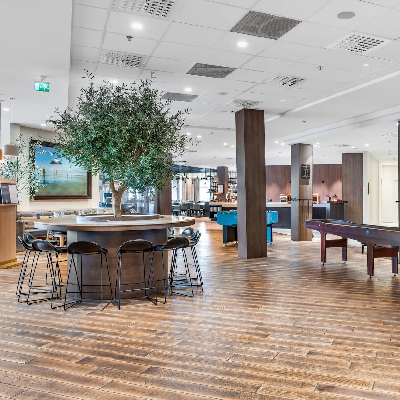 A spacious, modern interior with wooden floors, circular tables, black chairs, and olive trees. It's a well-lit, clean area, likely a lobby or lounge.