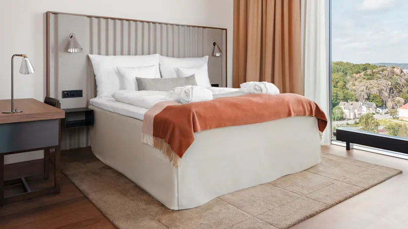 A neatly made bed with white linens and an orange throw sits in a modern, well-lit room. Beside it is a wooden desk, and a large window reveals a scenic view.