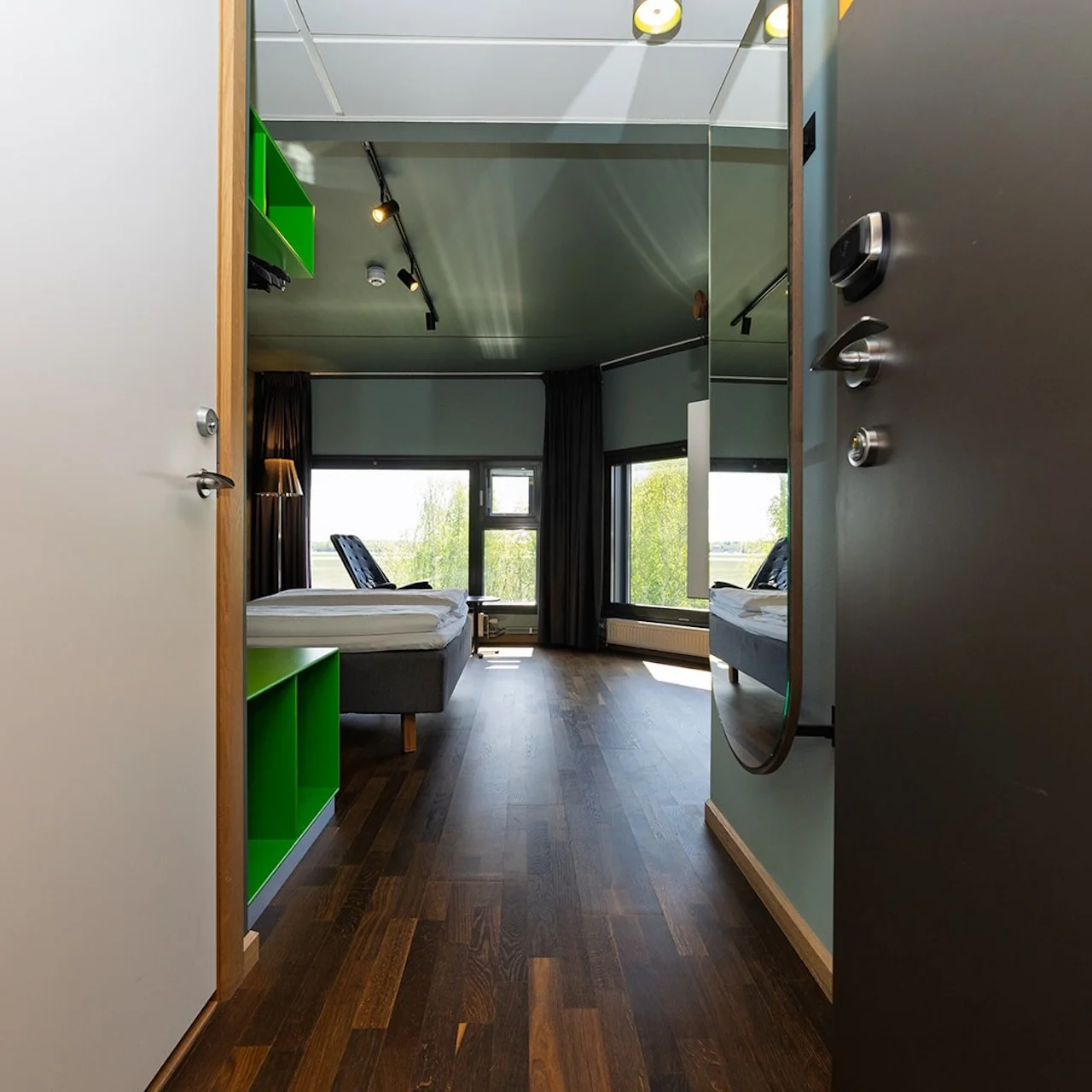 A room with two single beds, chairs, and a window view of greenery, seen through an open doorway with wooden flooring.
