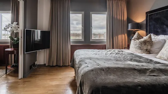 A cozy bedroom features a neatly made bed with pillows, situated beside two windows with cityscape views. A TV is mounted on the wall, and a lamp provides ambient lighting.