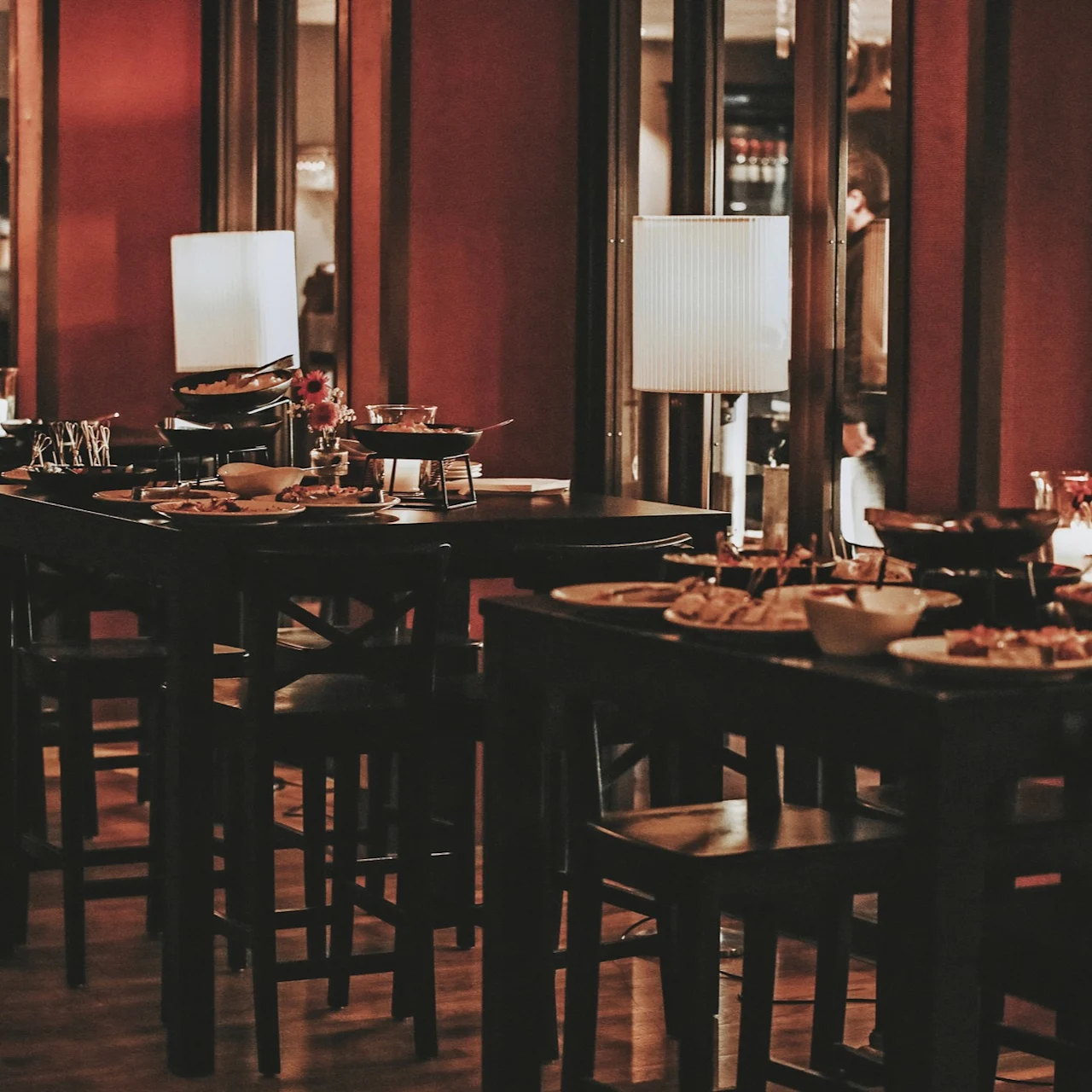 Tables adorned with plates and lamps in a dimly lit restaurant. Dishes and glasses are arranged neatly, surrounded by dark wooden furniture and red walls, creating a cozy ambiance.