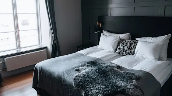 En pænt opredt seng med bløde puder og en mønstret pude, dækket af et gråt tæppe, står ved siden af et stort vindue med tunge gardiner i et svagt oplyst, moderne soveværelse.