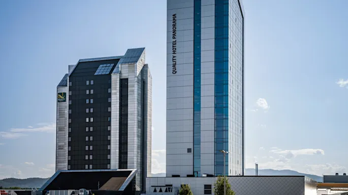 Kaksi korkeaa rakennusta seisoo lähekkäin kirkkaan sinisen taivaan alla. Yhdessä on pystysuuntainen teksti "QUALITY HOTEL PANORAMA". Kaupunkimaisemassa on matalia kukkuloita taustalla.