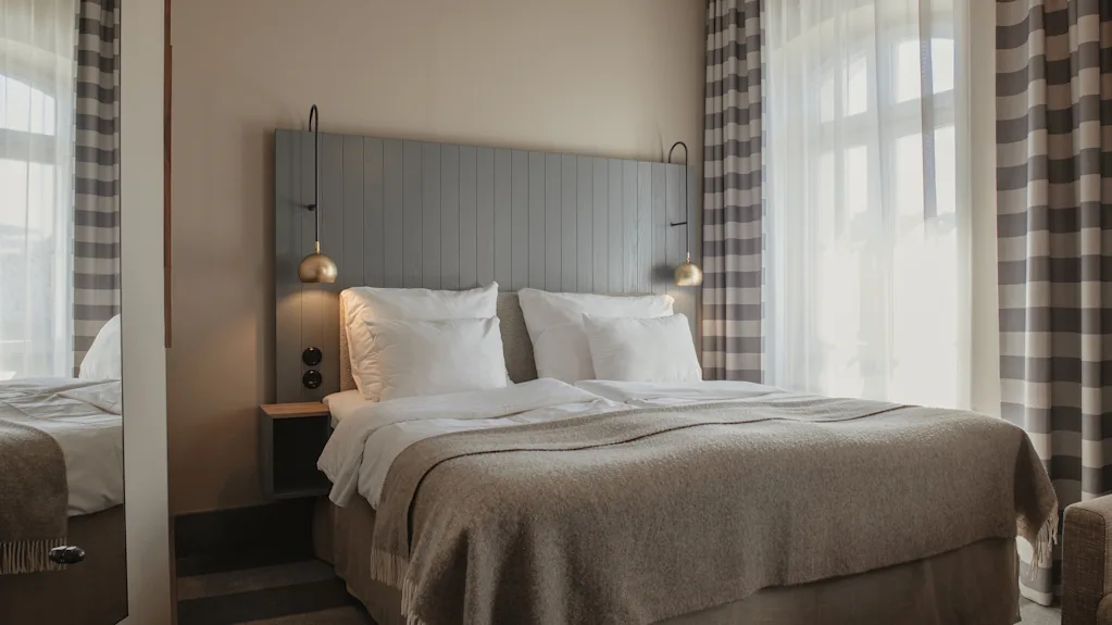 A cozy hotel room with a neatly made bed, bedside lamps, and striped curtains, ready for a comfortable stay.