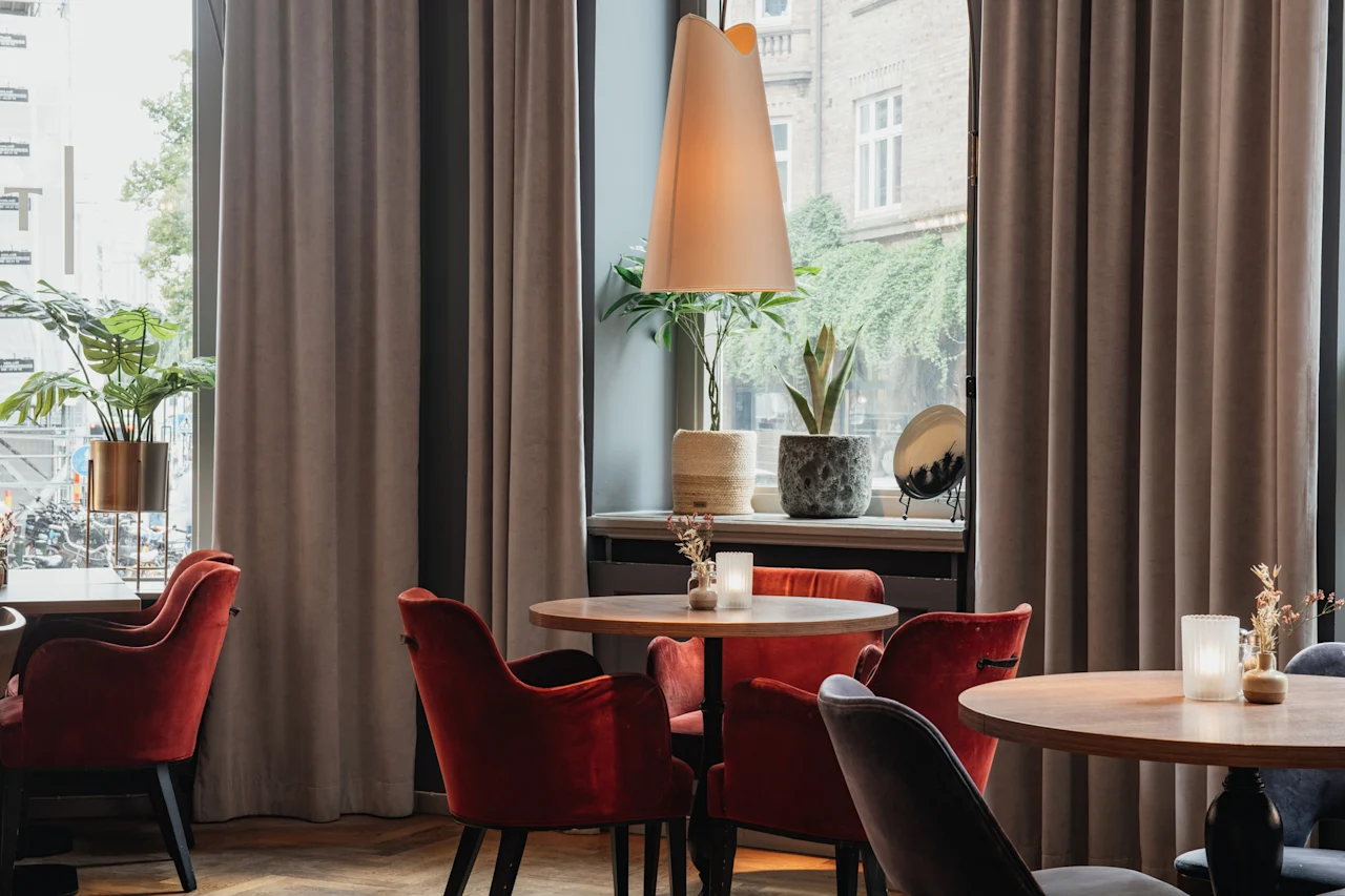 Et varmt og innbydende kafé- eller restaurantinteriør med runde trebord, plysj-røde stoler og planter ved store vinduer.