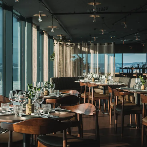 Bordene er dekket med vinglass og bestikk i en moderne restaurant, omgitt av store vinduer og myk belysning, som gir en naturskjønn utsikt.